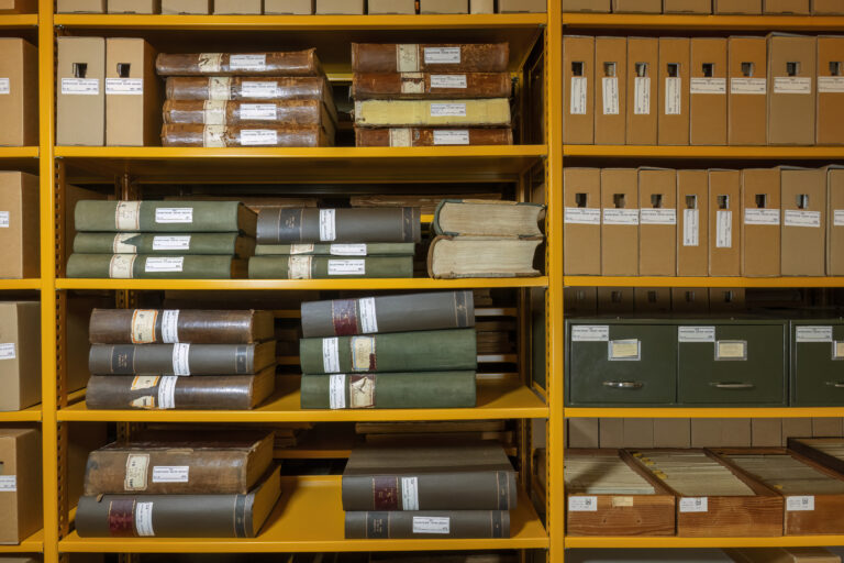 Een gele stellingkast met 5 planken aan de linkerkant met boeken erop. Aan de rechterkant ook 5 planken mat archiefdozen erop. Op de laatste plank staan 4 kaartenbakken.
