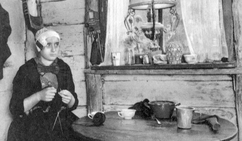 Een zwart wit foto met links een vrouw in klederdracht zittend aan een ronde tafel. De vrouw is aan het breien.
