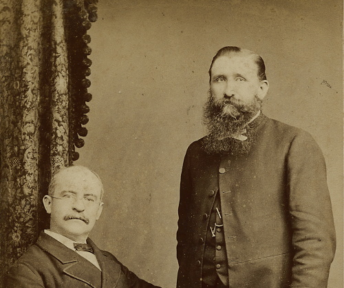 Foto in sepia kleur van twee manen. Rechts een zittende man en links een staande man.