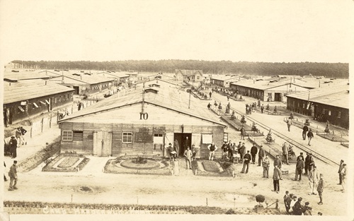 5900 Prentbriefkaarten Harderwijk – 492 Overzicht Belgen-Kamp Een prentbriefkaart met in het midden een loods waar nummer 10 opstaat. Aan de rechterkant en linkerkant staan gebouwen en daar staan ook mensen.