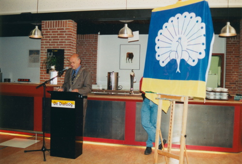Een kleurenfoto met aan de linkerkantde vlag van Ermelo gele strepen boven en onder. in het midden blauw met een witte pauw. Links staat een man achter een zwarte spreektafel.