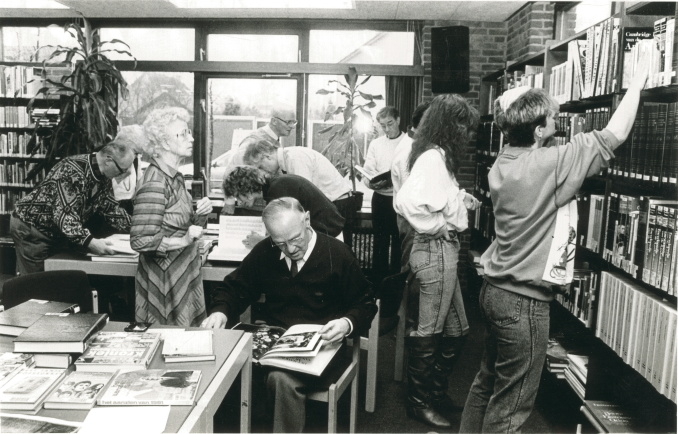 Verschillende mensen staan en zitten in een omgeving met boeken. Er staan twee tafels. En op de voorgrond een man die zittend door een boek aan het bladeren is.