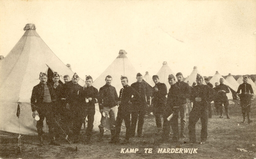 Op de Voorgrond een aantal mannen in uniform. Achter hun staan een aantal tenten. Als titel heeft deze prentbriefkaart Kamp te Harderwijk.