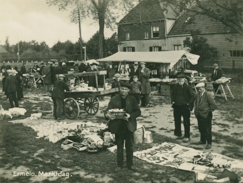 Zwart/wit foto met met mensen op een marktdag. Rechtsachterin staat een huis en voor het huis een kraampje.