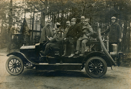 Een zwart/wit foto met zes mannen op een oude brandweerauto Linksachter staat een huis en achter de personen op de wagen staat een rij bomen.