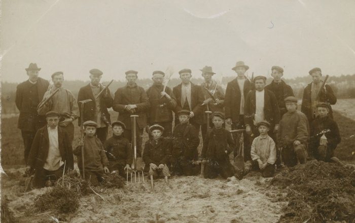 Een zwart/wit foto met op de achtergrond poserende mannen en op de voorgrond poserende kinderen op het land.
