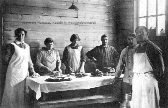 Zwart/wit foto met achter de tafel vier vrouwen staand. Rechts voor de tafel staan twee mannen.