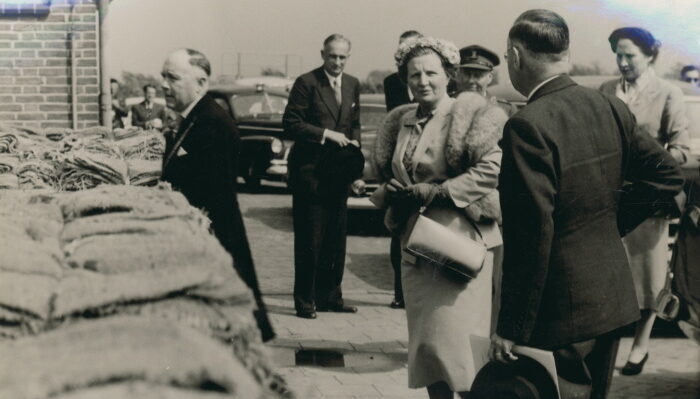 Zwart/wit foto van een groep mensen en koningin Juliana. aan de linkerkant liggen zandzakken.