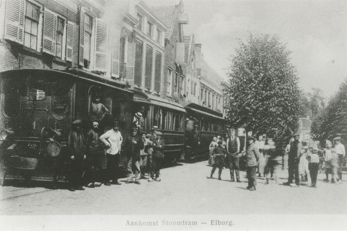 Zwart/wit foto van een tram, aan de linkerkant voor een rij huizen. Aan de rechterkant een groep mensen. Als tekst staat er: Aankomst Stoomtram - Elburg.